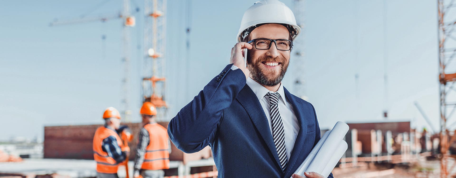 A smiling businessman in a suit and hard hat talking on a smartphone at a construction site, with workers and cranes in the background.